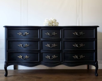 May include: A black dresser with nine drawers and ornate brass handles. The dresser has a curved front and legs, and a bouquet of white flowers sits on top. The furniture piece is painted in a matte black finish.