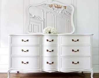 May include: A white dresser with nine drawers and ornate bronze handles. A matching white mirror with a decorative top is above the dresser. A bouquet of white flowers sits on the dresser's surface. The dresser has curved legs.