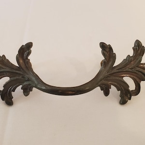 May include: Ornate bronze-toned drawer pull with a curved handle and stylized leaf design. The decorative hardware features a dark patina and is suitable for cabinets, dressers, or other furniture. The overall design is vintage.
