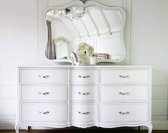 May include: A white dresser with nine drawers and ornate silver handles. Above the dresser is a matching white mirror with a decorative frame. The mirror reflects a chandelier and other decor, creating a bright and elegant bedroom furniture set.