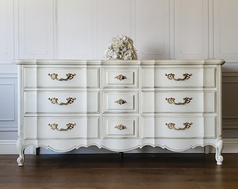 May include: A white dresser with nine drawers and ornate gold-colored handles. The dresser has a scalloped base and is topped with a bouquet of white flowers. The frame is accented with a subtle gray trim.