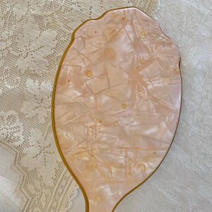 May include: A vintage hand mirror with a pink, marbled, plastic surface and a gold-colored frame.