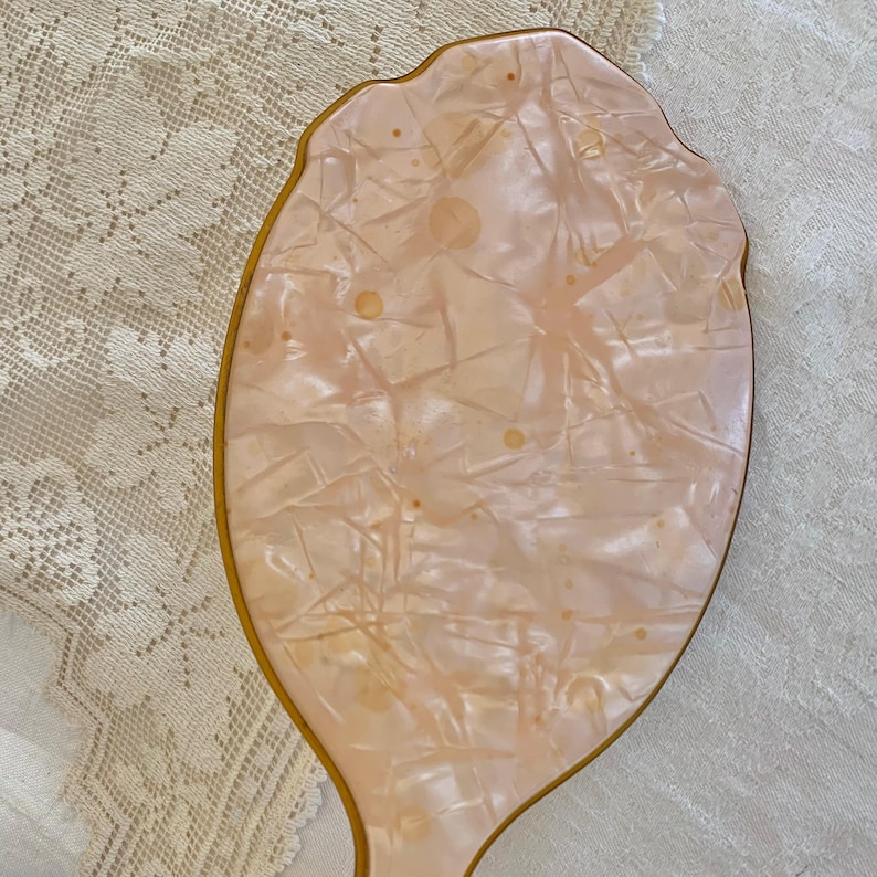 May include: A vintage hand mirror with a pink, marbled, plastic surface and a gold-colored frame.