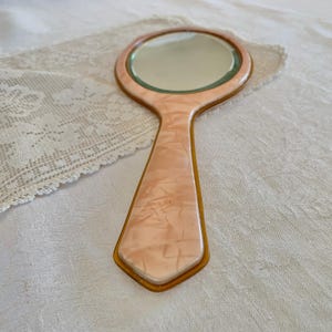 May include: A vintage hand mirror with a pink marbled plastic handle and a gold trim. The mirror has a round shape and is surrounded by a gold frame.