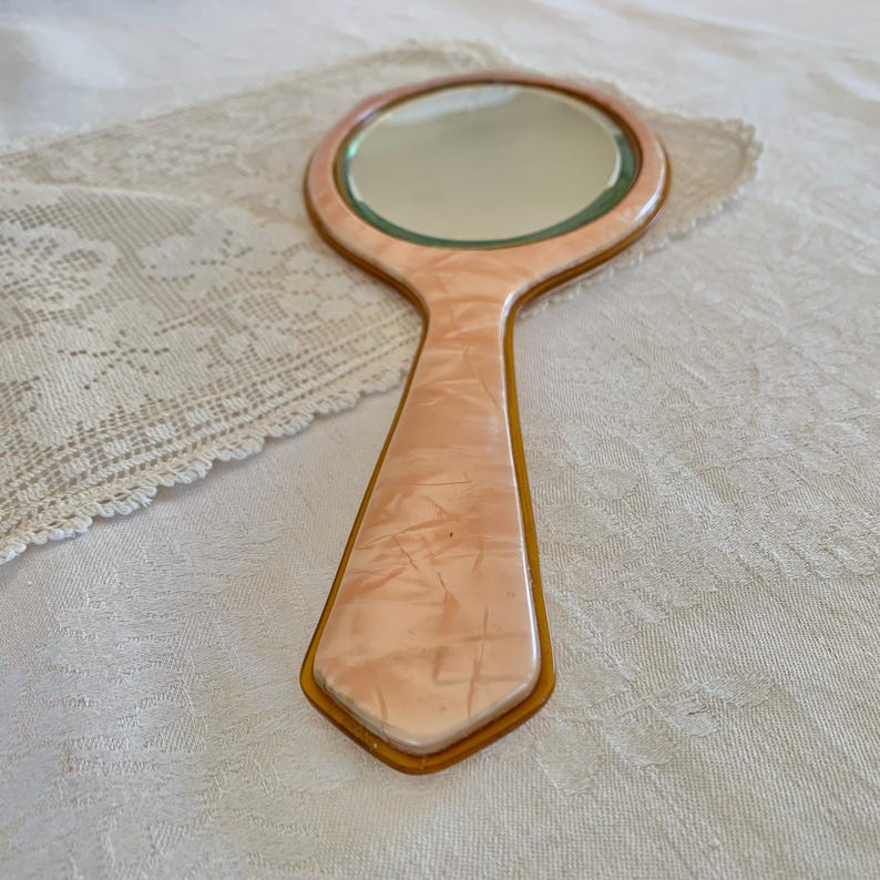 May include: A vintage hand mirror with a pink marbled plastic handle and a gold trim. The mirror has a round shape and is surrounded by a gold frame.