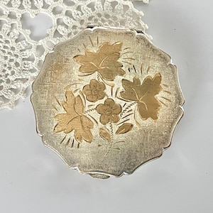 May include: Silver-toned compact with gold-colored floral design. The compact has a textured surface and a decorative leaf and flower pattern. The compact is round with a scalloped edge.