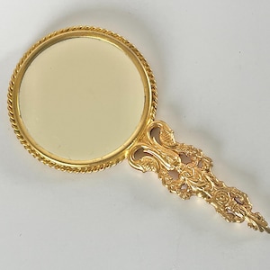 May include: A gold-coloured hand mirror with an ornate handle and a round mirror. The mirror's frame has a twisted rope design. The handle features a floral and scroll design. The mirror is likely vintage or antique.