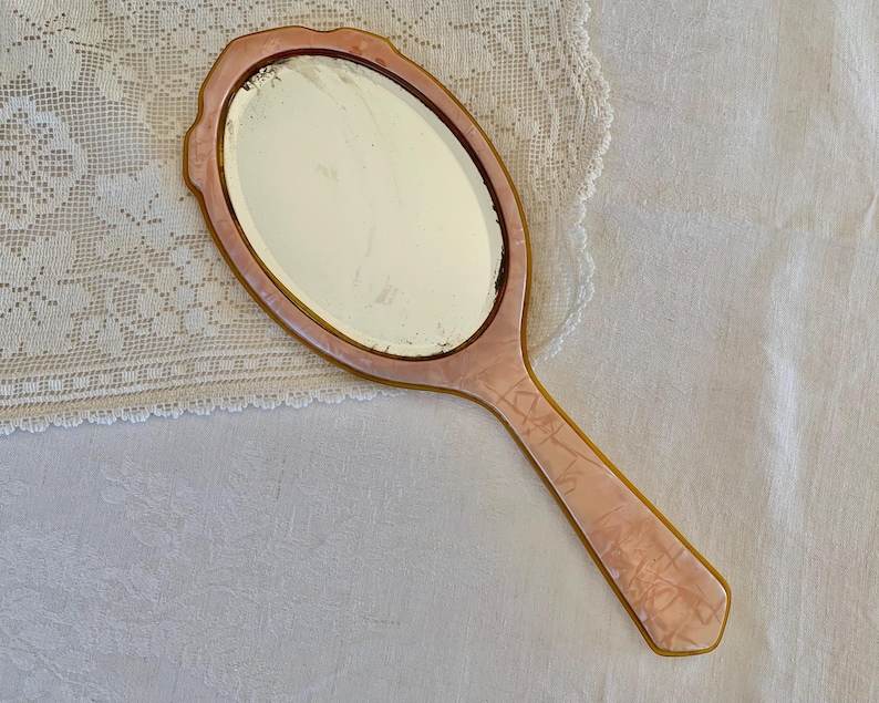 May include: A vintage hand mirror with a pink and white marbled plastic handle and a round, clear glass mirror.