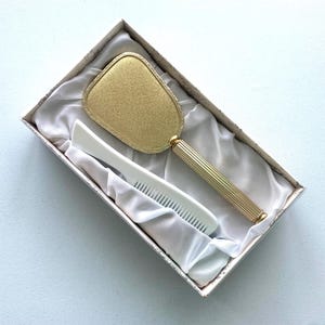 May include: A vintage vanity set in a box, featuring a gold-toned hand mirror and a white comb. The mirror has a rectangular shape with a gold handle. The comb is white with fine teeth. The set is presented on a white satin-like fabric.