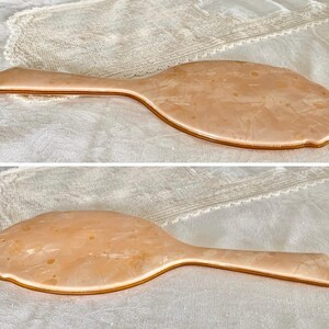 May include: A vintage hand mirror with a peach-colored plastic frame and a gold-colored trim. The mirror is oval shaped and has a slightly textured surface.