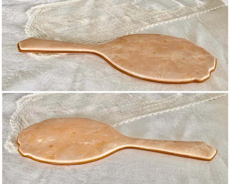 May include: A vintage hand mirror with a peach-colored plastic frame and a gold-colored trim. The mirror is oval shaped and has a slightly textured surface.