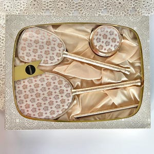 Puede incluir: Un juego de tocador decorativo en una caja con borde dorado. El conjunto incluye dos espejos de mano, un estuche compacto y un palito, todos con un estampado floral sobre un fondo crema. Los mangos y los bordes son dorados. La caja está forrada con satén.