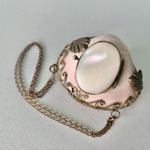 以下が含まれることがあります： 真珠光沢のある楕円形の中心部と繊細なブロンズチェーンが付いた、ヴィンテージの貝殻型ペンダント。ペンダントはピンクがかった色合いで、装飾的なブロンズのディテールとアクセントが施されています。