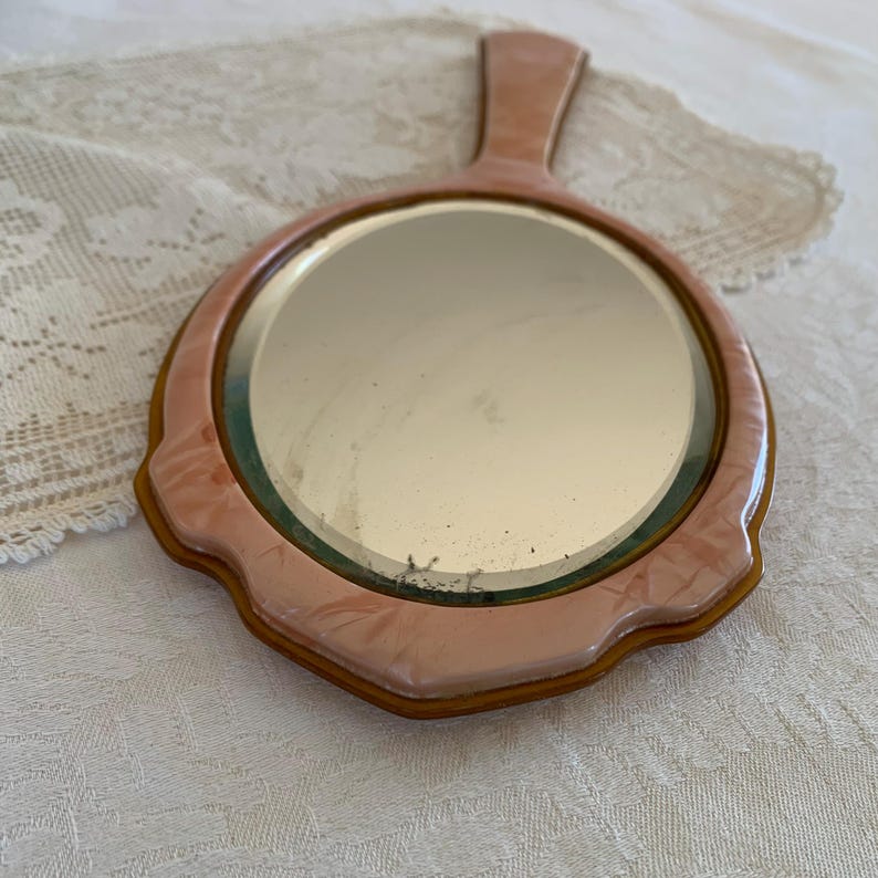 May include: A vintage hand mirror with a pink plastic frame and a gold trim. The mirror has a round shape and a handle.
