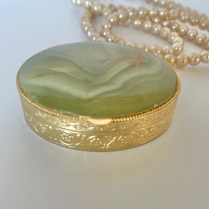 May include: A small, round, gold-coloured trinket box with a light green, marbled stone top. The box has an ornate, embossed design around the sides. A strand of faux pearls is in the background.