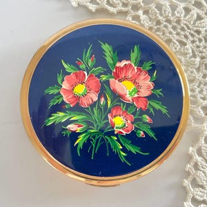 Puede incluir: Un estuche compacto redondo y vintage con una tapa azul marino con un diseño floral. Las flores son rojas con centros amarillos y hojas verdes. El estuche compacto tiene un borde de metal dorado.