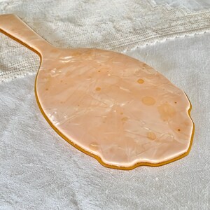 May include: A vintage hand mirror with a peach-colored plastic handle and a peach-colored reflective surface. The mirror has a scalloped edge and a swirl pattern.