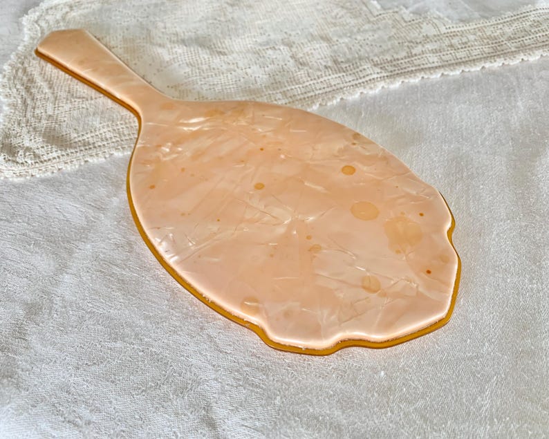May include: A vintage hand mirror with a peach-colored plastic handle and a peach-colored reflective surface. The mirror has a scalloped edge and a swirl pattern.