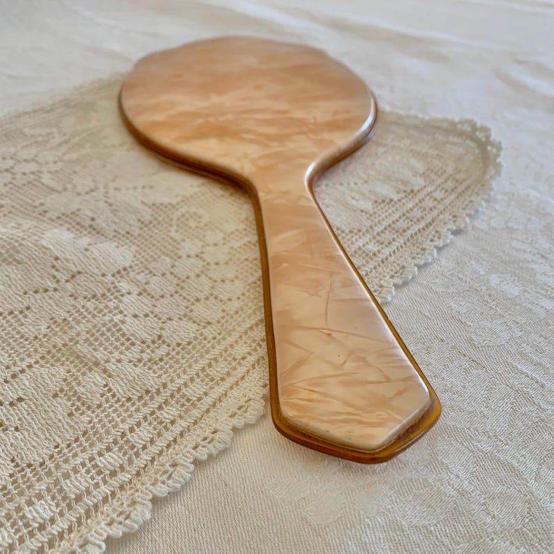 May include: A vintage hand mirror with a peach-colored plastic handle and a round mirror. The handle has a marbled pattern.