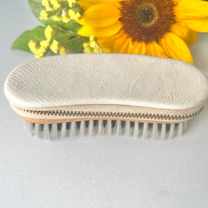 May include: A cream-colored, oval-shaped nail brush with a zippered leather case. The brush has a wooden base and white bristles. A yellow sunflower and small yellow flowers are in the background.