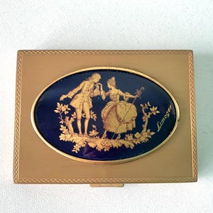 May include: A rectangular gold-toned compact with an oval, dark blue center. The center features a gold-colored illustration of a couple in period clothing, with the word "Limoges" in gold. The compact has a decorative border.