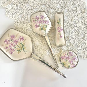 Peut inclure: Un ensemble de coiffeuse vintage comprenant un miroir à main, une brosse, un poudrier et un petit objet rectangulaire. Chaque pièce présente une surface crème avec des fleurs violettes brodées et des accents verts, dans des cadres argentés.