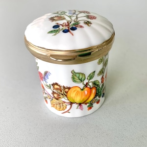 May include: A white porcelain trinket box with a lid. The box is decorated with colorful floral and fruit designs, including apples and berries. The lid has a gold-colored metal rim.