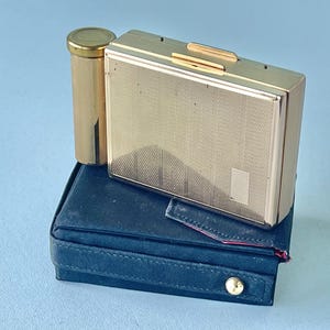 May include: A gold-toned compact, lipstick tube, and a dark blue case. The compact has a textured surface and a rectangular shape. The lipstick tube is cylindrical with a matching gold cap. The case is rectangular with a button closure.