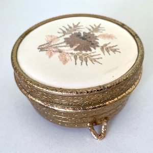 Puede incluir: Una caja de baratijas vintage dorada con un diseño floral bordado en la tapa. La caja tiene forma redonda, detalles ornamentados y patas pequeñas. La tapa presenta una tela color crema con bordados marrones y rosas.