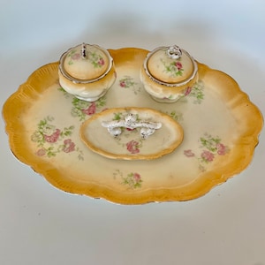 May include: A vintage porcelain vanity set with a tray, two lidded jars, and a small dish with a decorative handle. The set features a floral pattern with pink roses and green leaves on a cream background.
