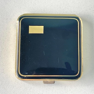 May include: A vintage compact mirror, square in shape. It features a navy blue surface with a gold rectangular accent and a gold border. The compact has a gold-coloured clasp.
