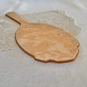 May include: A vintage hand mirror with a peach-colored plastic frame and a swirl pattern. The mirror has a scalloped edge and a handle.