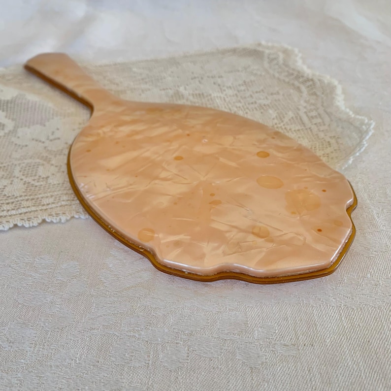May include: A vintage hand mirror with a peach-colored plastic frame and a swirl pattern. The mirror has a scalloped edge and a handle.