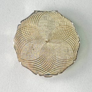 Puede incluir: Un espejo compacto plateado vintage con un borde festoneado único. La superficie presenta un intrincado diseño grabado de líneas curvas superpuestas que irradian desde un punto central. El compacto está cerrado y parece estar en buenas condiciones.