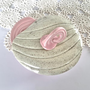 May include: A ceramic trinket dish with a speckled, off-white base and a pink accent. The dish has a rounded shape with a textured surface and a decorative pink loop. The dish is likely used for holding small items.