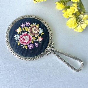 May include: A vintage hand mirror with a silver handle and frame. The round mirror has a black fabric backing with an embroidered floral design in pink, yellow, and purple. The handle has a twisted rope design.