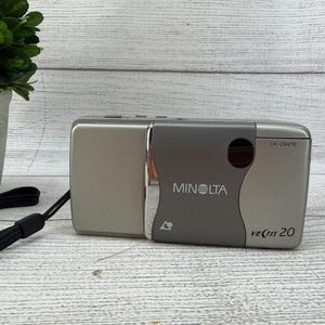 May include: A silver and grey Minolta Vectis 20 IX-Date compact film camera with a black wrist strap. The camera body features a metallic finish, a dark grey front panel with the Minolta logo, a circular lens cover, and the model name printed in black.