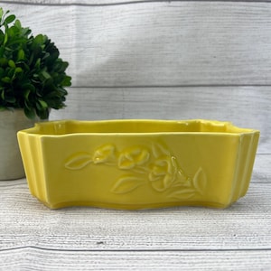 May include: A yellow ceramic planter with a floral design. The planter has a rectangular shape with rounded edges and a slightly wavy rim.