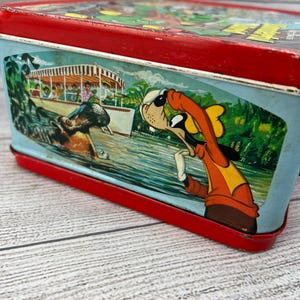 Op de afbeelding: Vintage metalen lunchbox met een rood frame en een kleurrijke illustratie. De illustratie toont een stripfiguur in een oranje outfit, een boot met passagiers en een nijlpaard in het water. De lunchbox heeft een blauwe achtergrond.