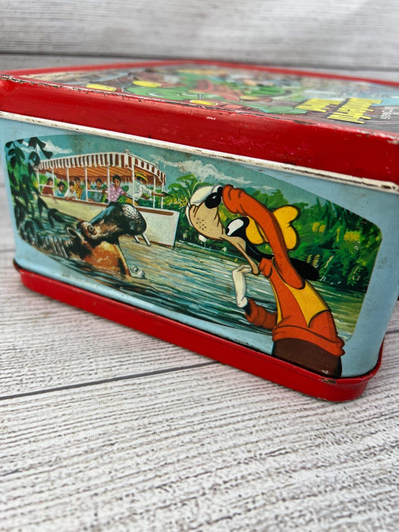 Op de afbeelding: Vintage metalen lunchbox met een rood frame en een kleurrijke illustratie. De illustratie toont een stripfiguur in een oranje outfit, een boot met passagiers en een nijlpaard in het water. De lunchbox heeft een blauwe achtergrond.