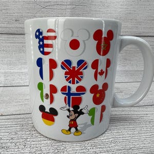 Puede incluir: Taza de cer&aacute;mica blanca con asa. La taza presenta un dise&ntilde;o de Mickey Mouse con banderas de diferentes pa&iacute;ses del mundo. Las banderas incluyen Estados Unidos, Jap&oacute;n, Reino Unido, Canad&aacute;, M&eacute;xico y China.