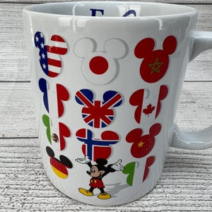 Puede incluir: Taza de cer&aacute;mica blanca con Mickey Mouse y banderas de diferentes pa&iacute;ses del mundo. La taza tiene un asa en el lado derecho. Las banderas incluyen Estados Unidos, Jap&oacute;n, Marruecos, Reino Unido, Canad&aacute;, M&eacute;xico, Finlandia, China, Alemania e Italia.