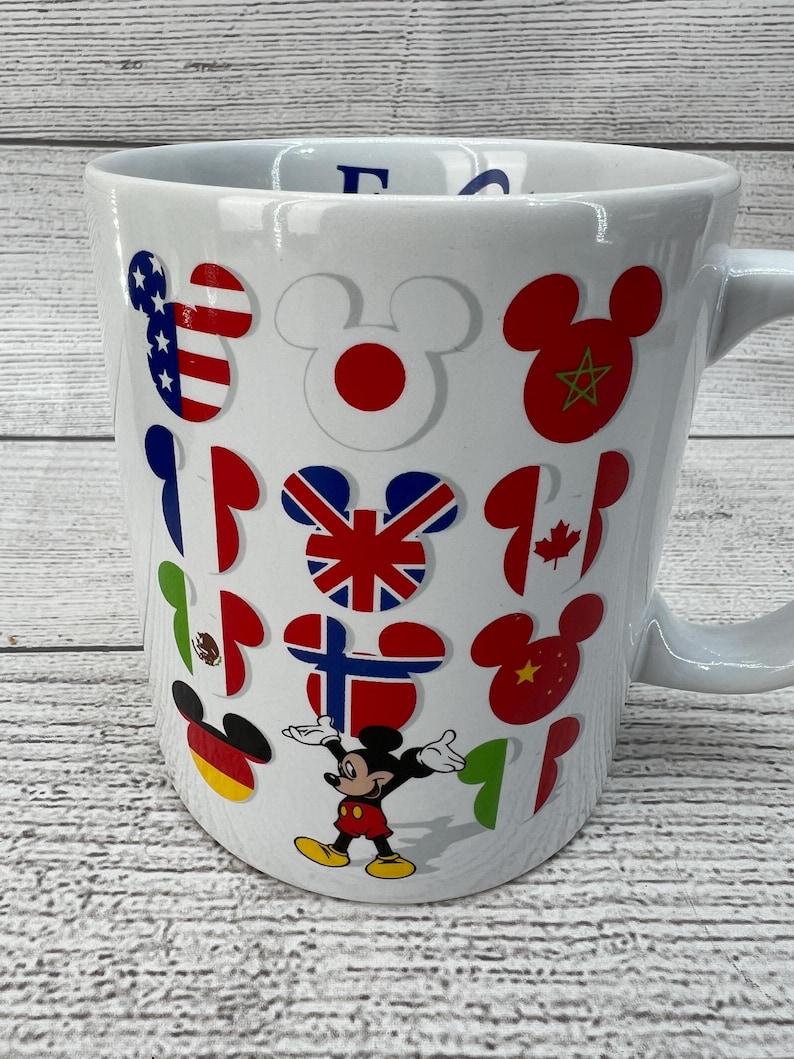 Puede incluir: Taza de cer&aacute;mica blanca con Mickey Mouse y banderas de diferentes pa&iacute;ses del mundo. La taza tiene un asa en el lado derecho. Las banderas incluyen Estados Unidos, Jap&oacute;n, Marruecos, Reino Unido, Canad&aacute;, M&eacute;xico, Finlandia, China, Alemania e Italia.