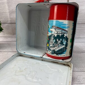 Op de afbeelding: Vintage metalen lunchbox met een rode handgreep en een bijpassende rode thermoskan. De thermoskan heeft een kleurrijke illustratie van een futuristische trein en mensen. De lunchbox is lichtblauw met een wit interieur.
