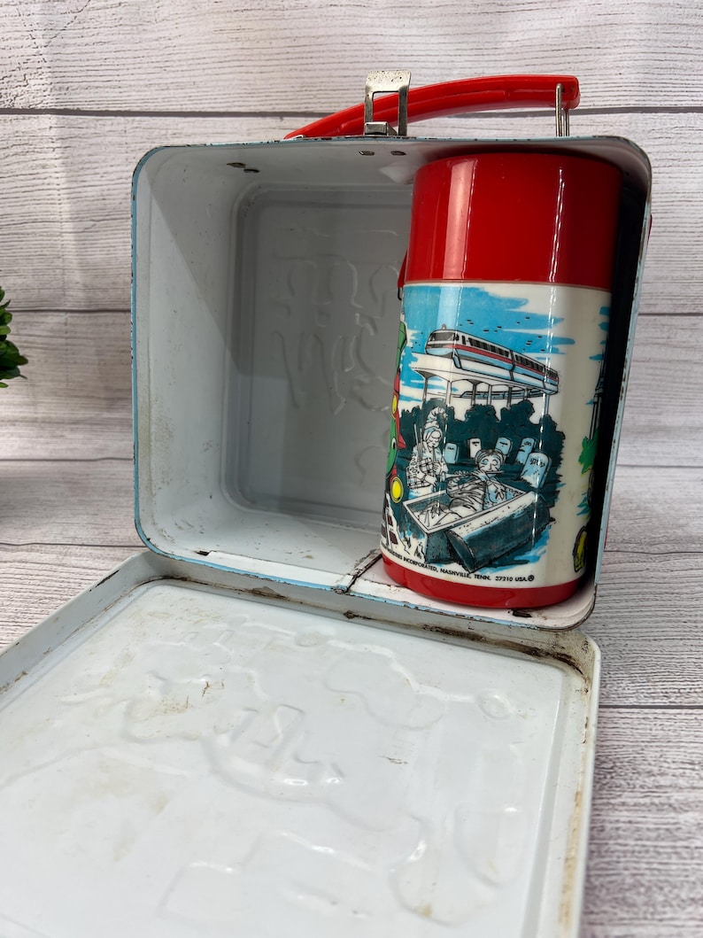 Op de afbeelding: Vintage metalen lunchbox met een rode handgreep en een bijpassende rode thermoskan. De thermoskan heeft een kleurrijke illustratie van een futuristische trein en mensen. De lunchbox is lichtblauw met een wit interieur.