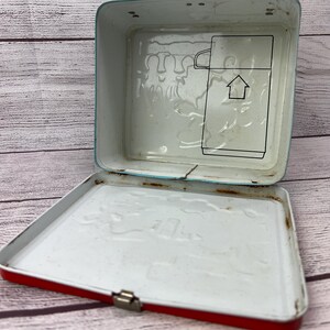 May include: Red metal lunchbox with a white interior. The lid has a raised design of a house and people. The bottom of the lunchbox has a raised design of a house and people.