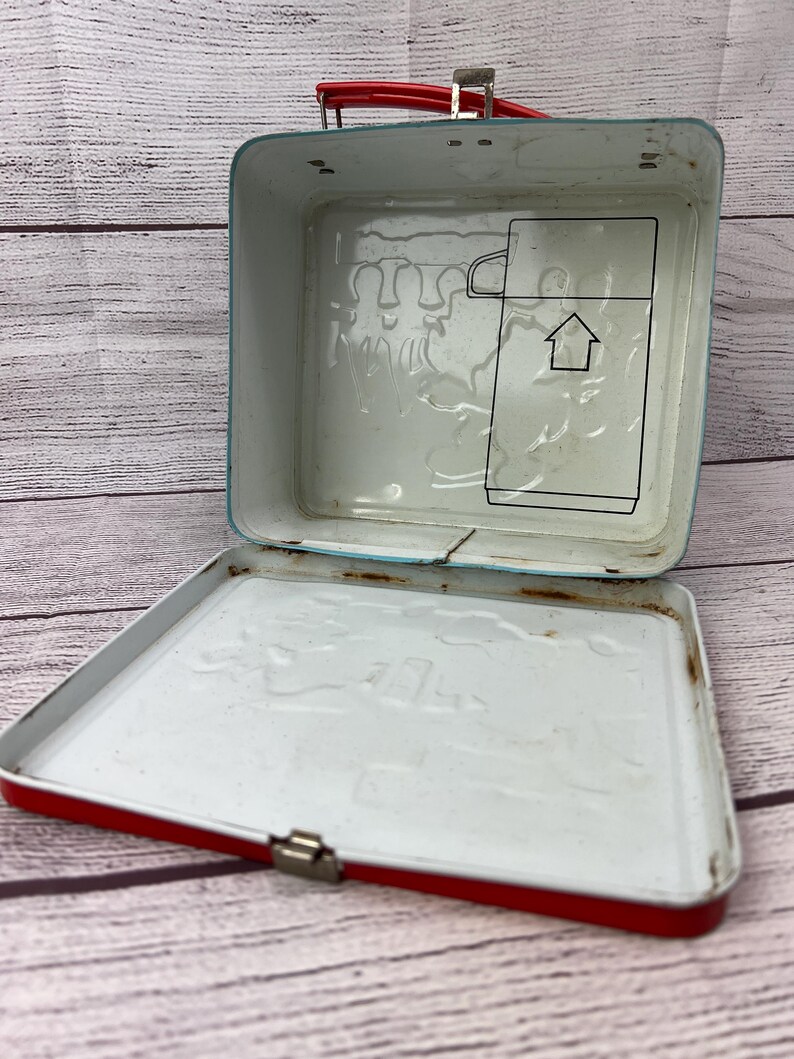 May include: Red metal lunchbox with a white interior. The lid has a raised design of a house and people. The bottom of the lunchbox has a raised design of a house and people.