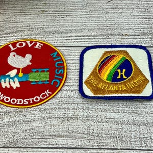 May include: Two embroidered patches. One is round, red, with a white dove on a guitar, and the words "PEACE LOVE MUSIC WOODSTOCK" in yellow. The other is rectangular, with a gold emblem, a rainbow, and the words "THE ATLANTA HILTON" on a white background.