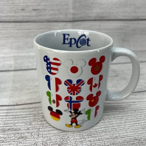 Puede incluir: Taza de cer&aacute;mica blanca con un logotipo "Epcot" azul y dise&ntilde;os de Mickey Mouse con banderas de diferentes pa&iacute;ses.