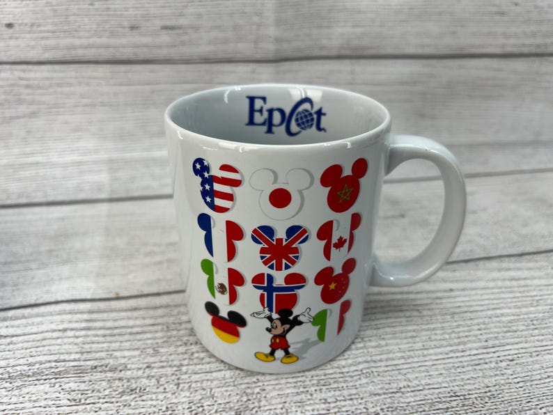 Puede incluir: Taza de cer&aacute;mica blanca con un logotipo "Epcot" azul y dise&ntilde;os de Mickey Mouse con banderas de diferentes pa&iacute;ses.